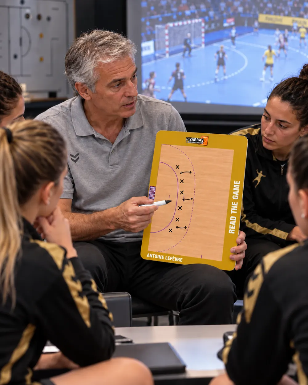 Ein Handballtrainer erklärt seinen Spielerinnen vor dem Videobildschirm einen Spielzug an der Taktiktafel